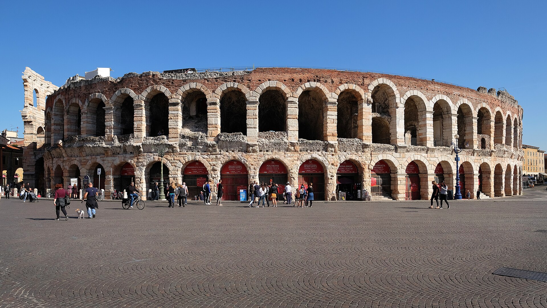 Esterno dell’Arena di Verona in piazza Bra, arcate e portoni rossi, passanti in piazza.